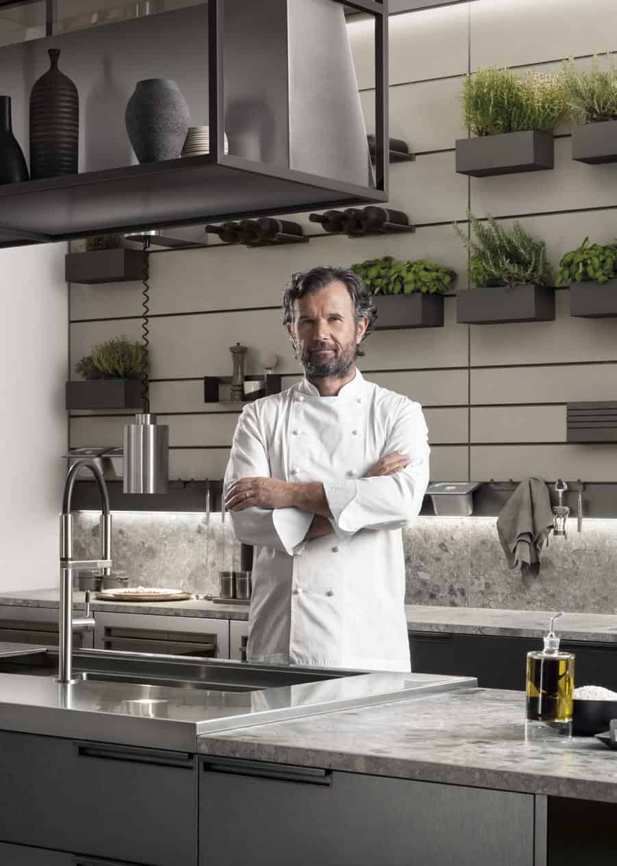 Chef standing in a modern kitchen with arms crossed, surrounded by herbs and kitchenware.