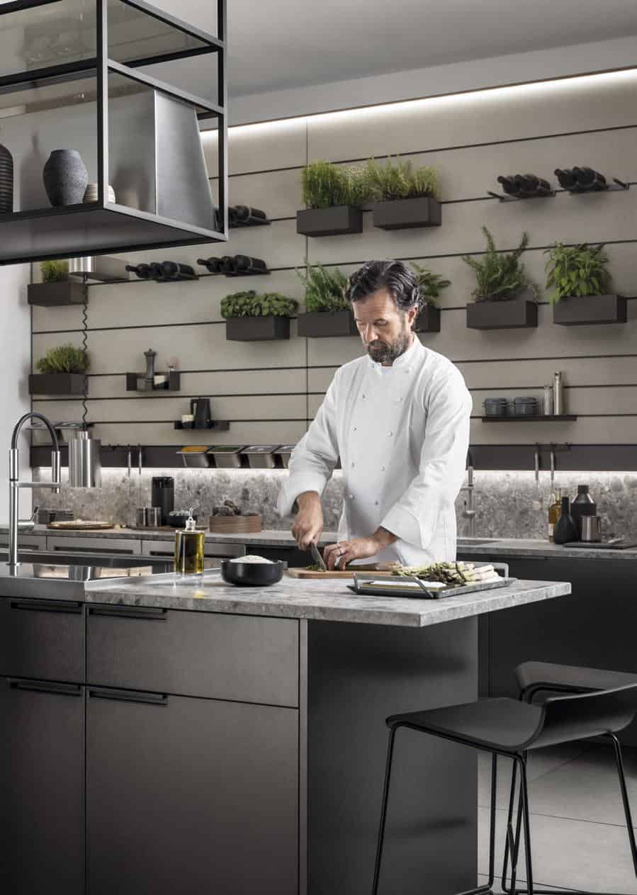 Chef in a modern kitchen preparing food on a marble island with shelves of plants and kitchen items in the background.