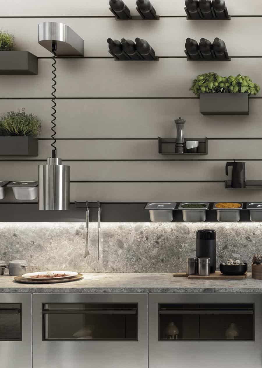 Modern kitchen interior with stainless steel pendant light, herb planters, shelves with bottles, spices, and a countertop featuring containers and utensils against a stone backsplash.