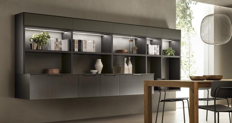 Wall-mounted shelving unit with cabinets, displaying books, plants, and decorative items, beside a wooden dining table and chairs, with a white pendant light above.