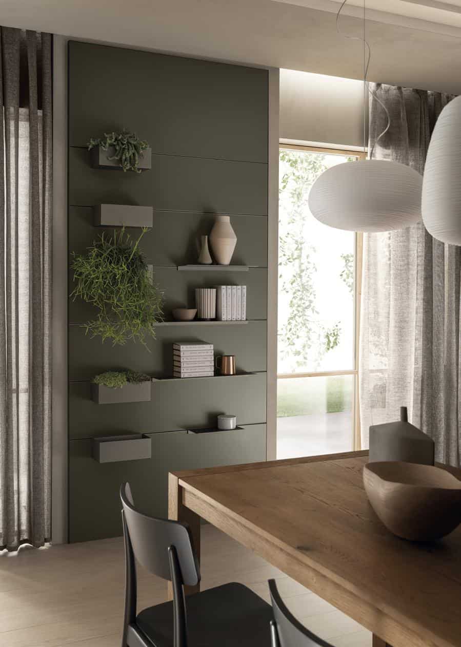 Modern interior with a wooden dining table, black chair, and wall-mounted shelves displaying books, plants, and vases; large windows with sheer curtains and hanging pendant lights.