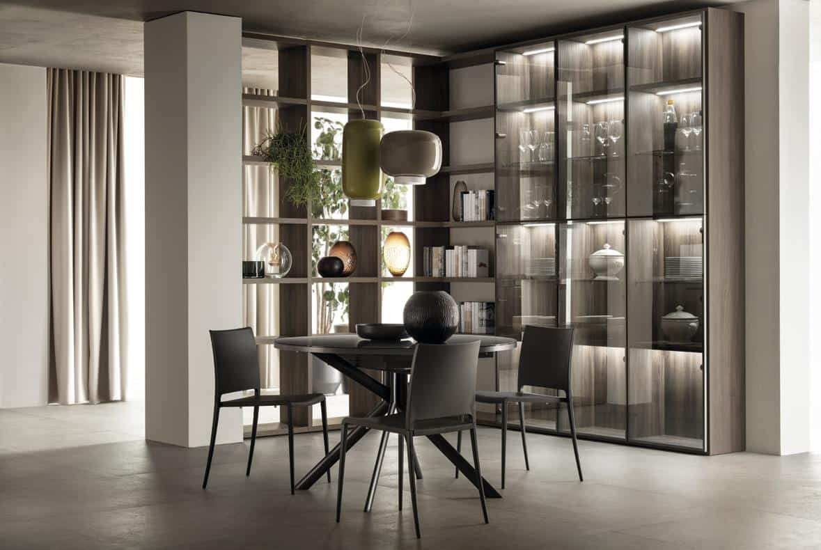 Modern dining room with a round table, four chairs, glass cabinet with dishware, open shelving with books and decor, and pendant lights.