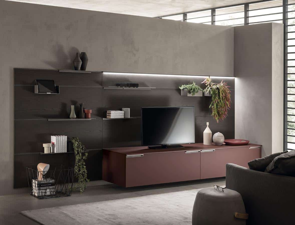 Modern living room with a TV on a sleek brown cabinet, wall-mounted shelves with decorative items, plants, a basket with magazines, and a gray sofa.