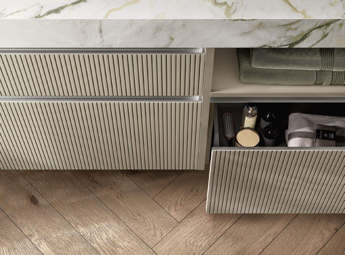 A modern bathroom vanity with ribbed cabinet panels, a marble countertop, and partially open drawers displaying toiletries and towels, set on a herringbone wood floor.