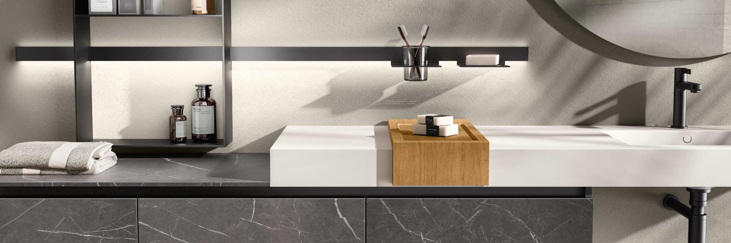 Modern bathroom vanity with a black faucet, white countertop, wooden tray, and wall-mounted shelves holding toiletries and towels.