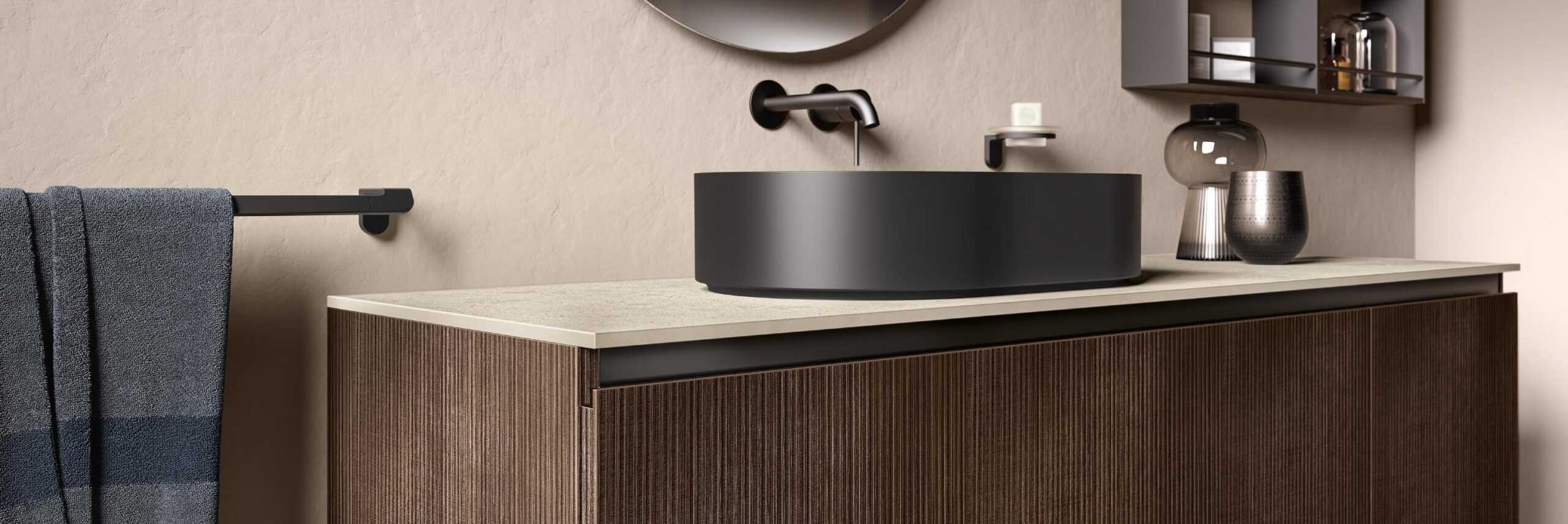 Modern bathroom with a textured dark wood vanity, black round vessel sink, wall-mounted faucet, gray towel, and shelves with decorative items.