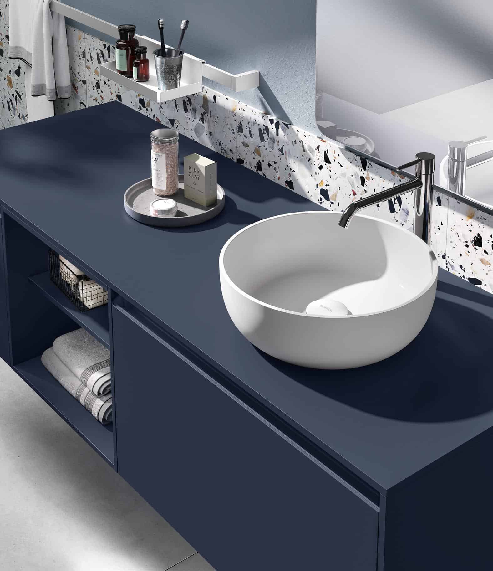 Modern bathroom vanity with a deep blue countertop, a round white vessel sink, a silver faucet, and shelving under the counter. Accessories like a tray with bath items are present, with terrazzo tile backsplash.