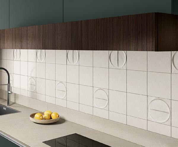 Kitchen backsplash with beige tiles featuring circular embossed patterns, under dark wood cabinets, with a bowl of lemons next to a sink and black cooktop.