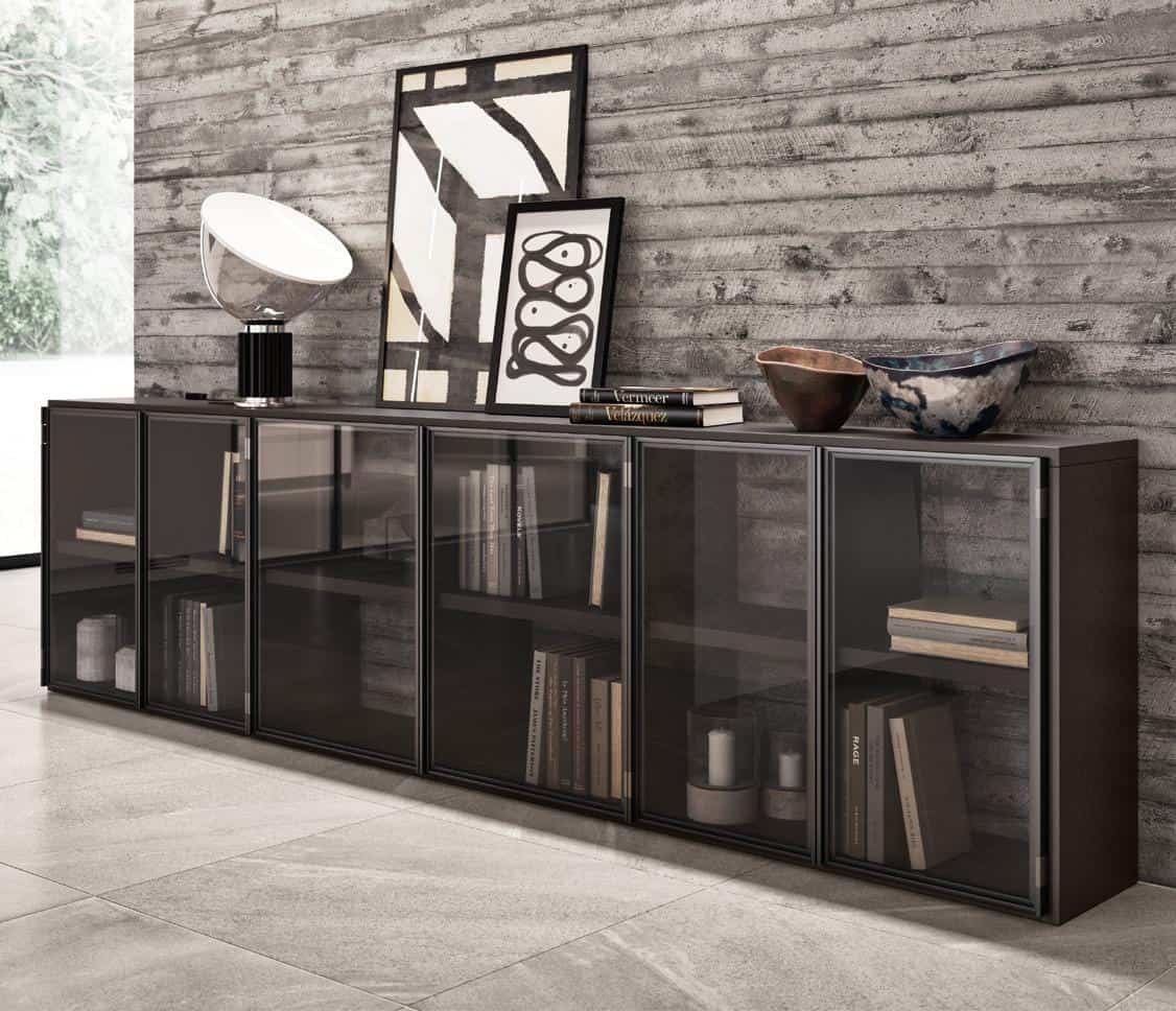 Modern interior with a low black cabinet featuring glass doors, displaying books and decorative items; a textured concrete wall, abstract art, a lamp, and ceramic bowls decorate the top.