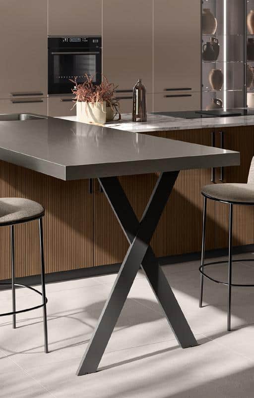 Modern kitchen with a sleek island, featuring a dark countertop and crossed metal legs, two stools, cabinets, oven, and decorative items.