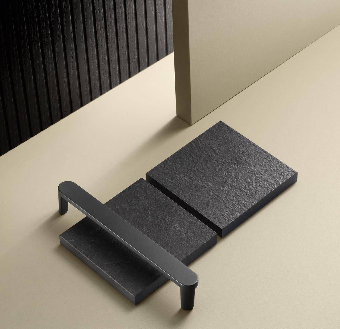 A minimalist black handle resting on two black textured square tiles on a beige surface.