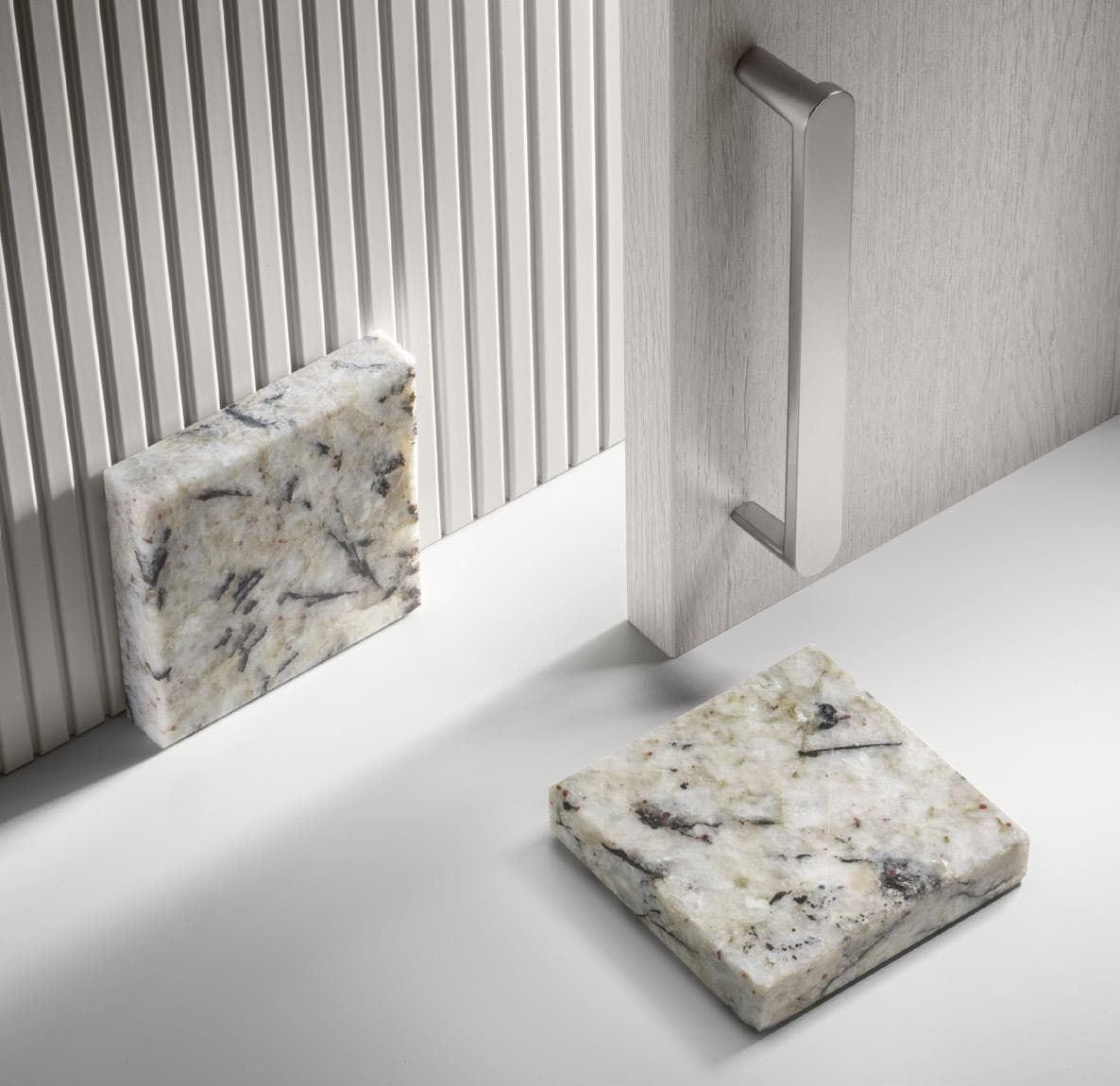 Two square marble tiles, one leaning against a ribbed wall and another on a flat surface, near a white wooden panel with a metal handle.