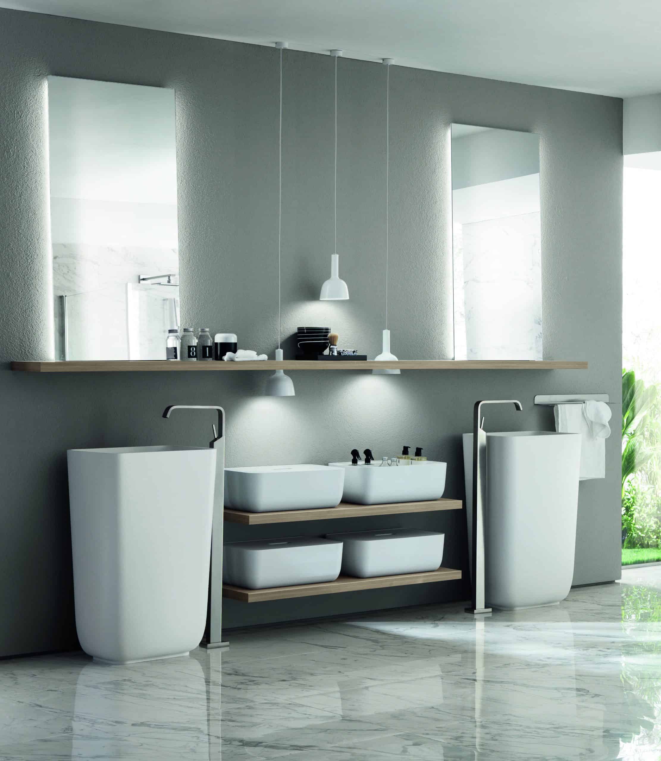 Modern bathroom interior with two tall sinks, a shelf with toiletries, pendant lights, and large mirrors, all set against a gray wall with marble flooring.