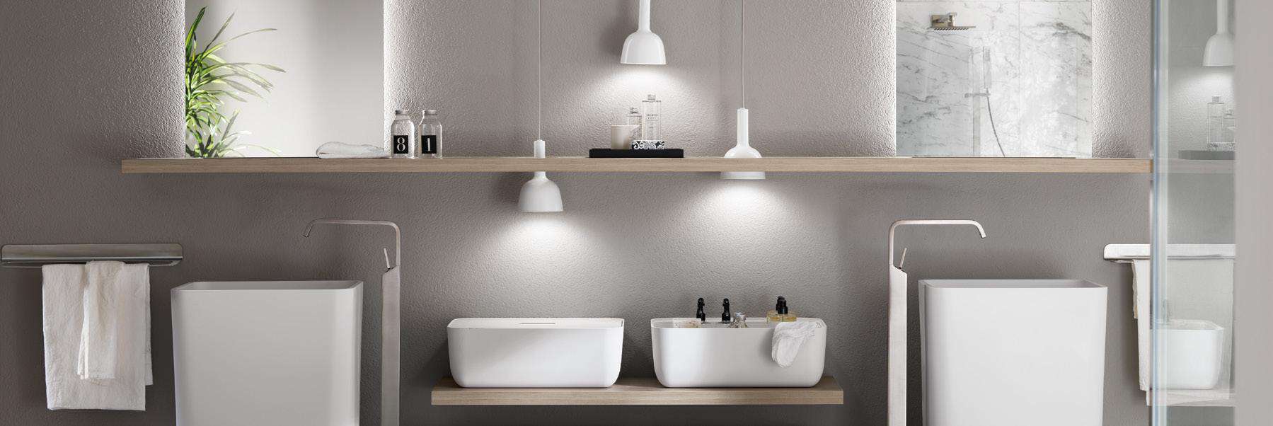 Modern bathroom with two white rectangular basins on a light wood shelf, minimalist fixtures, and soft lighting from two pendant lamps, set against a gray wall.
