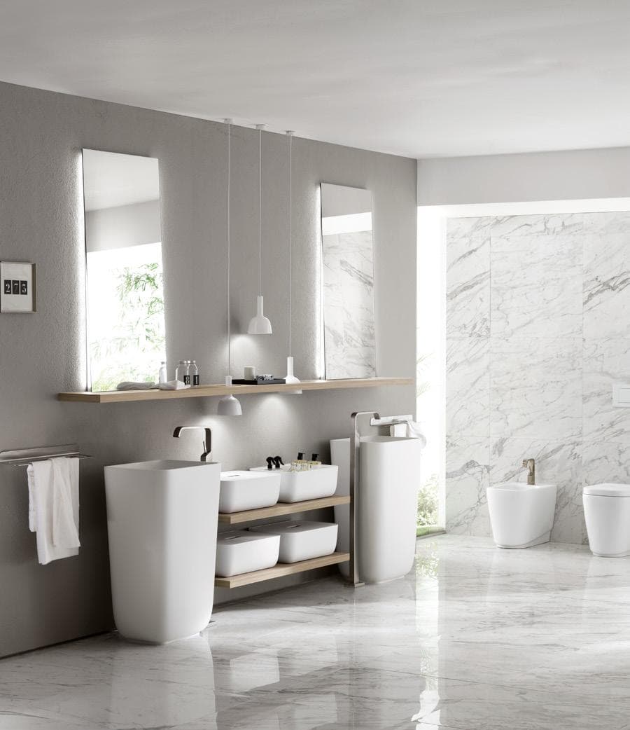 Modern bathroom with two pedestal sinks, a wooden shelf, large mirrors, pendant lights, a towel rack, and marble flooring and walls.