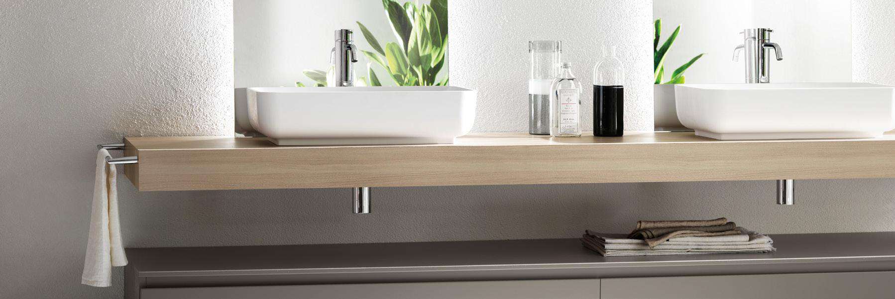 Modern bathroom with two white vessel sinks on a wooden countertop, chrome faucets, decorative bottles, and folded towels beneath.