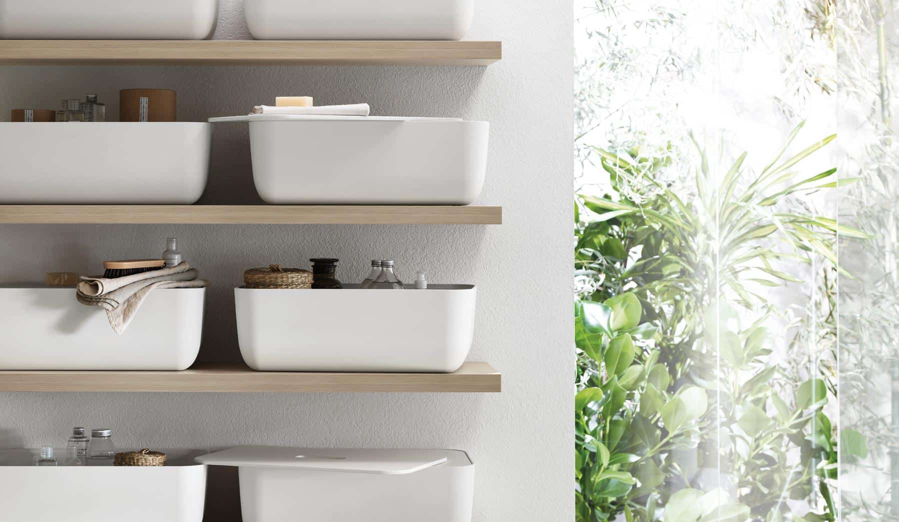White storage bins on wooden shelves with assorted toiletries, next to a large window with green foliage outside.