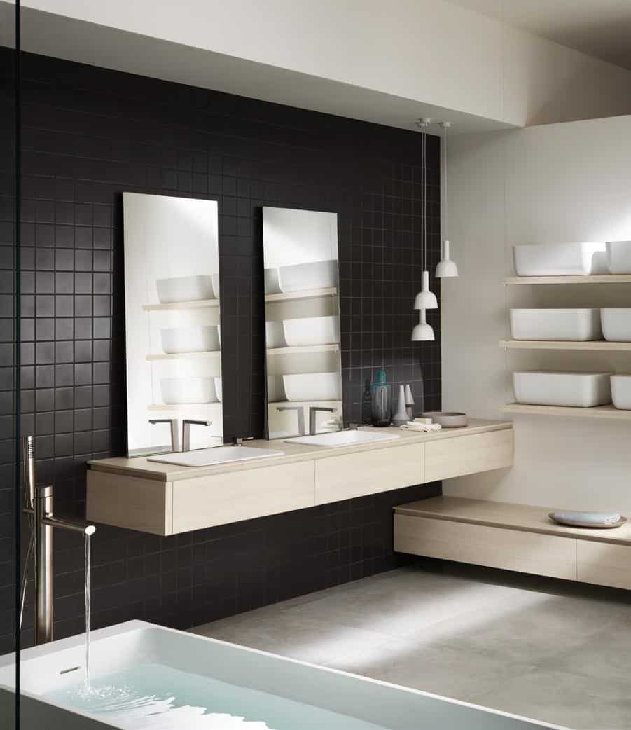 Modern bathroom with black tiled wall, dual mirrors and sinks, wooden floating shelves, and a filling bathtub.