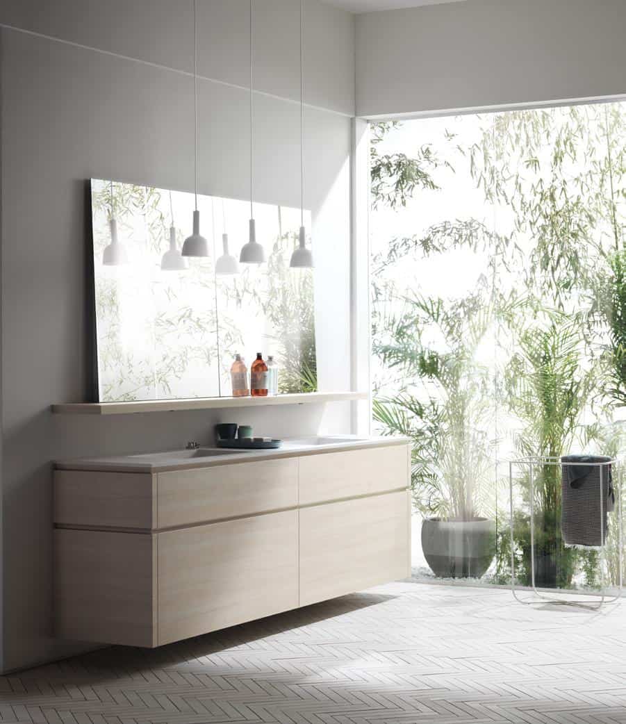 Modern minimalist bathroom with light wood cabinetry, large mirror, hanging pendant lights, and window view of lush greenery.
