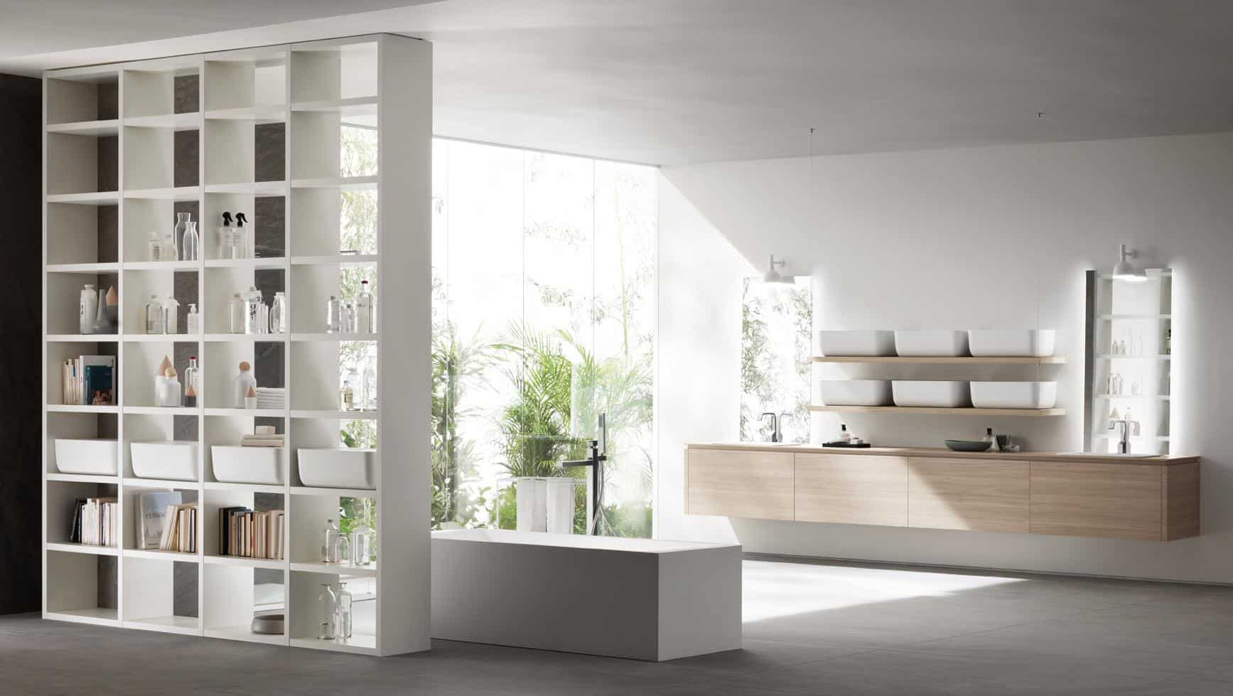 Modern bathroom with a white freestanding bathtub, open shelving filled with books and toiletries, a wall-mounted wooden vanity with rectangular sinks, large windows, and indoor plants.