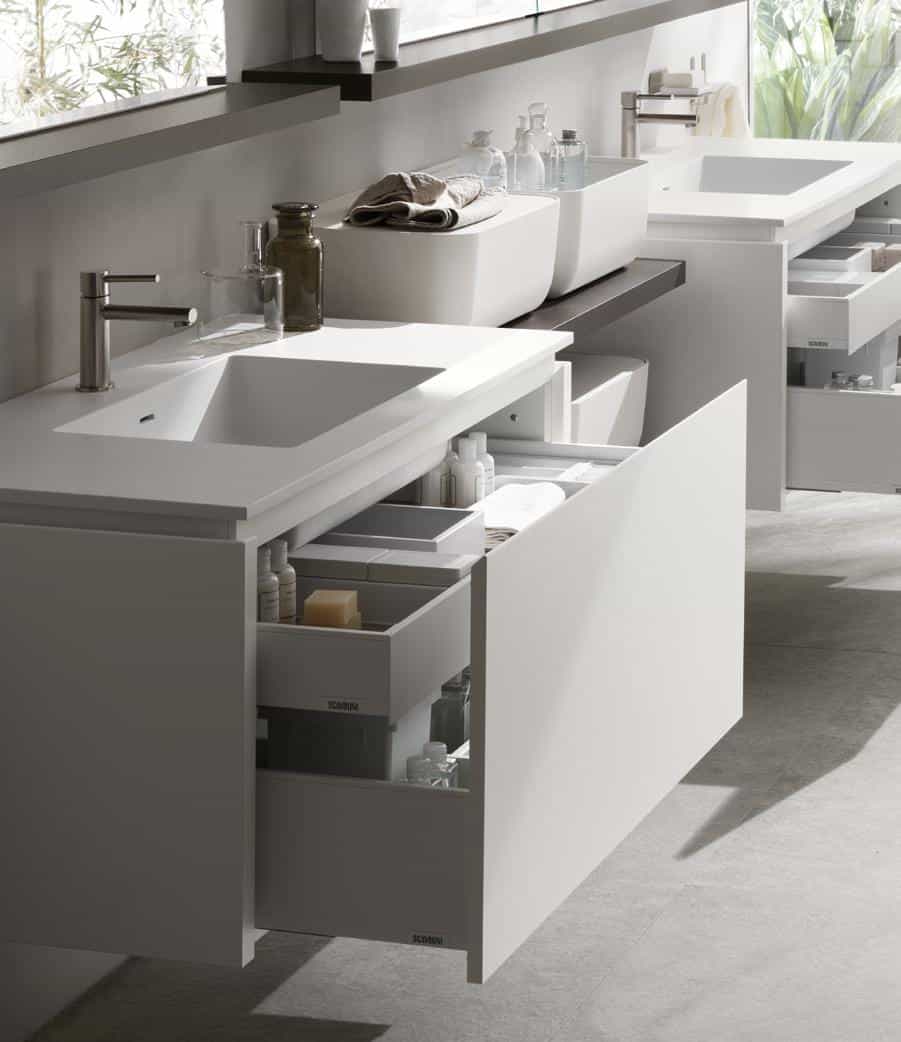 Modern bathroom with white vanity, open drawers containing toiletries, and two sinks with chrome faucets.