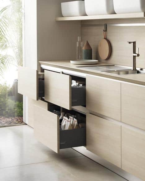 Modern kitchen with light wood cabinets, open drawers, and a stainless steel sink, featuring minimalist decor and a cutting board on the counter.