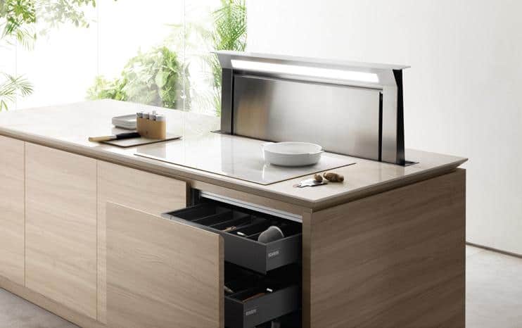 Modern kitchen island with an integrated downdraft ventilation system, open drawer, light wood cabinetry, and a bright, plant-filled background.