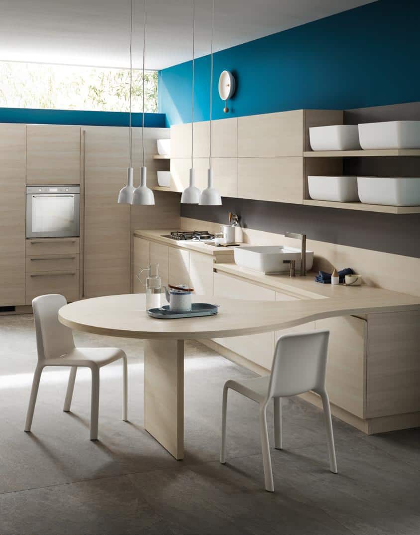Modern kitchen with light wood cabinets, a round table with two white chairs, hanging pendant lights, and a teal accent wall.