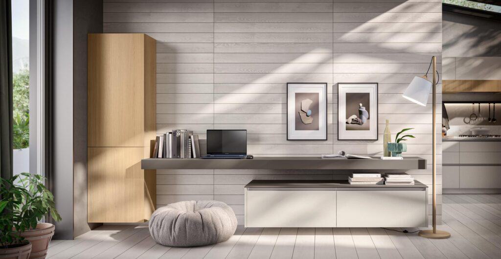 Modern home workspace with a wooden cabinet, wall-mounted desk holding a laptop and books, white floor lamp, cozy round pouf, and framed abstract art on a light wood-paneled wall.