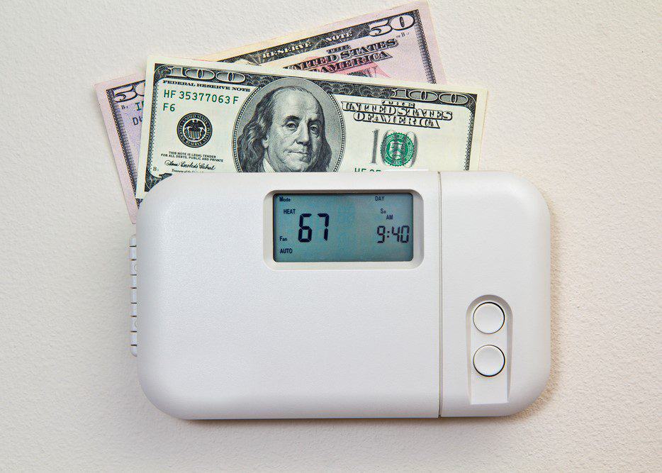 A digital thermostat set to 67 degrees Fahrenheit with two U.S. dollar bills tucked behind it, one fifty and one hundred.