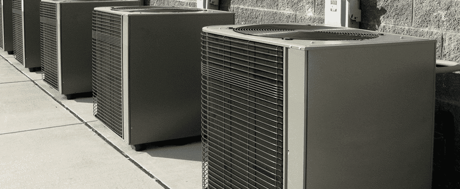A row of large outdoor air conditioning units is placed along a concrete walkway beside a textured stone wall.