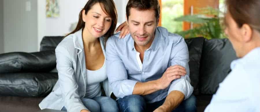 A couple sits on a couch, attentively listening to a third person in a bright room.