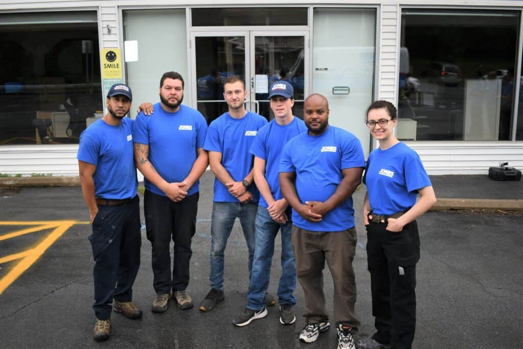 Six people in matching blue shirts and caps stand in front of a storefront with reflective windows.