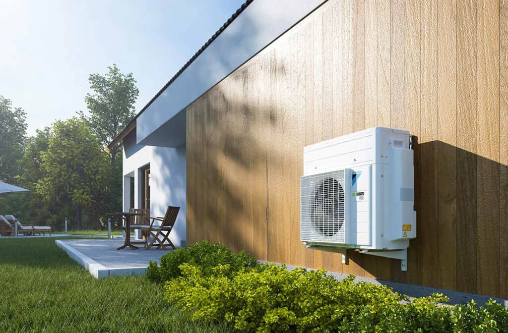Outdoor air conditioning unit mounted on a wooden exterior wall of a modern house, surrounded by green lawn and bushes, with patio furniture and trees in the background.