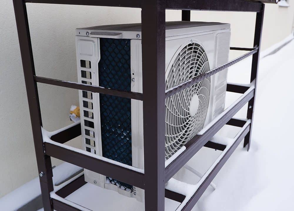 An outdoor air conditioning unit covered with a protective metal frame sits on the ground surrounded by snow.