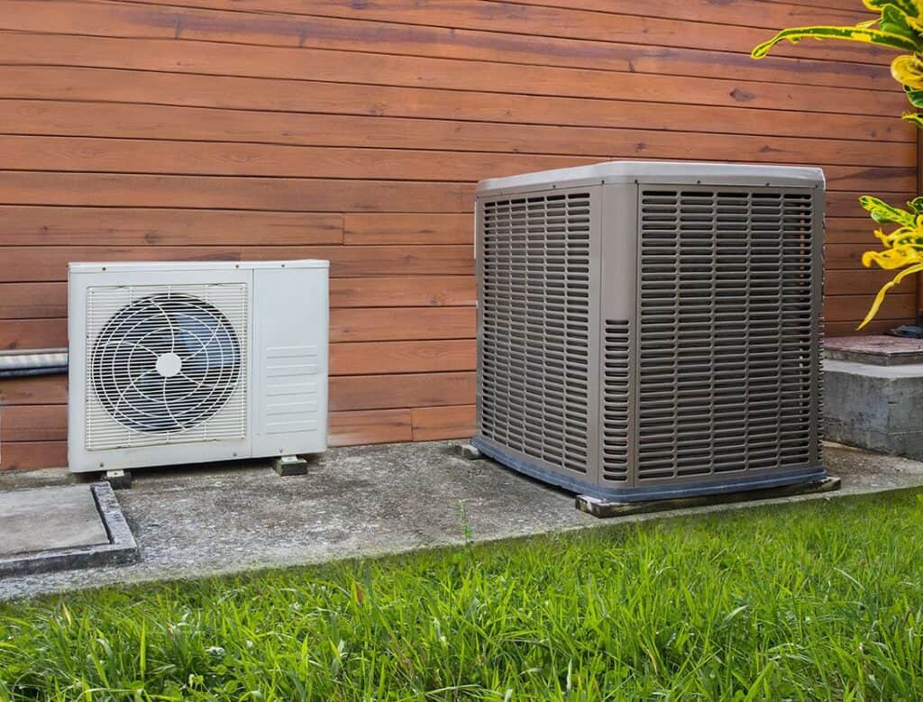 Two air conditioning units are installed outside on a concrete platform next to a wooden wall, surrounded by grass and plants.