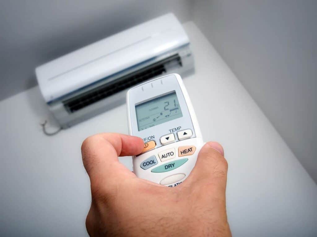 Hand holding a remote control aimed at a wall-mounted air conditioner. The remote shows a temperature setting of 21 degrees Celsius and various mode options.