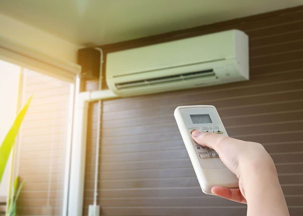 Hand holding a remote control pointed at a wall-mounted air conditioning unit in a room with wooden wall panels.