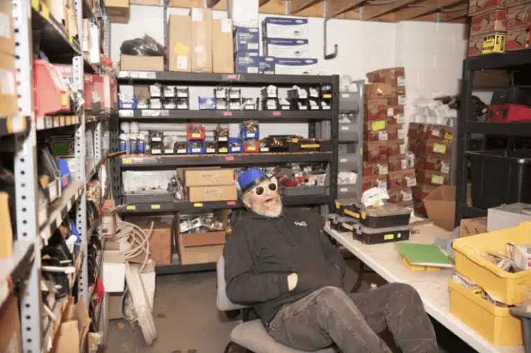 A person wearing sunglasses and a party hat sleeps on an office chair in a cluttered storage room with shelves full of boxes and various items.