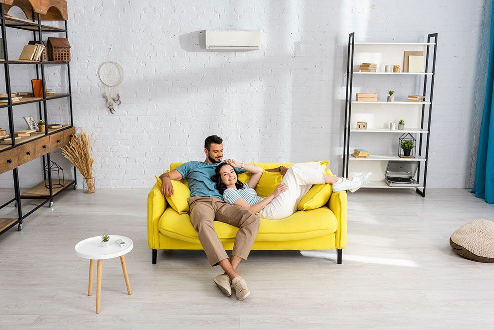 Couple relaxing on a bright yellow sofa in a modern living room with white walls, shelves, and minimal decor.