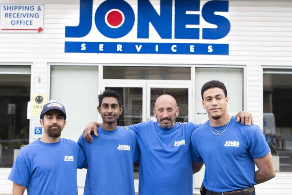 Four men in blue "Jones Services" shirts stand smiling in front of a company building with a "JONES SERVICES" sign above.