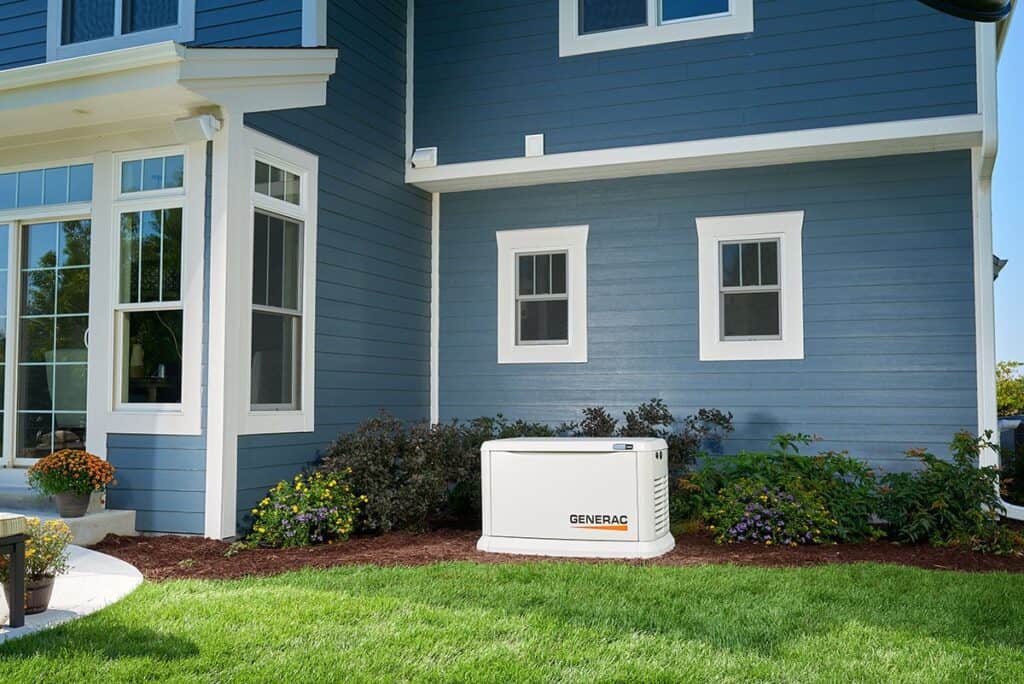 A Generac standby generator is installed outside a blue house, surrounded by landscaped plants and grass—showcasing how Mega Trends in HVAC technology are shaping modern home power solutions.