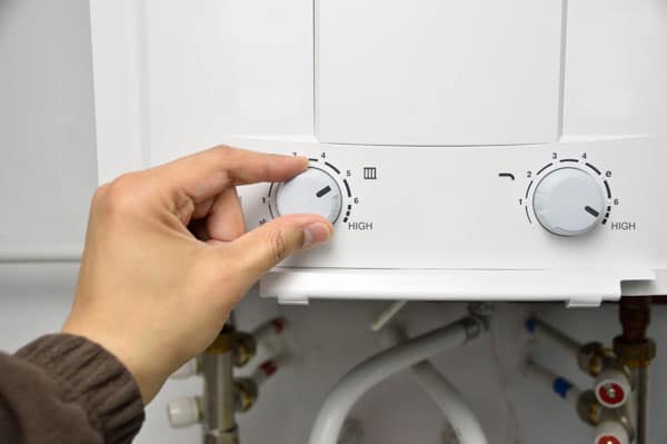 A person adjusts a temperature control knob on a white water heater unit with visible pipes below.