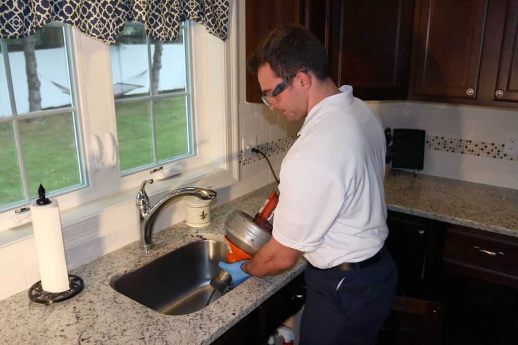 A person wearing gloves is using a plumbing snake to unclog a kitchen sink drain in a home kitchen.
