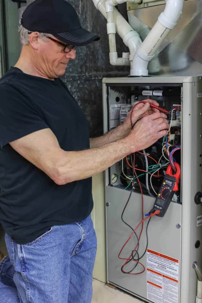 Man in black shirt and cap uses a multimeter for wiring diagnostics on an open HVAC unit.