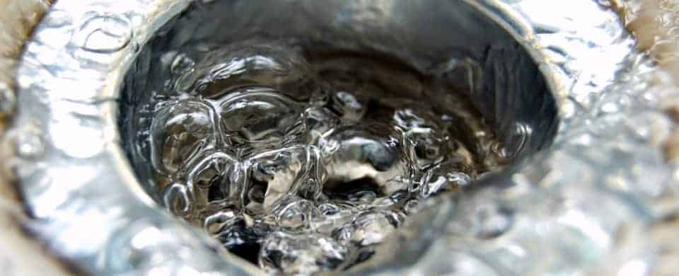 Close-up of water swirling down a metal sink drain, creating bubbles and reflections.