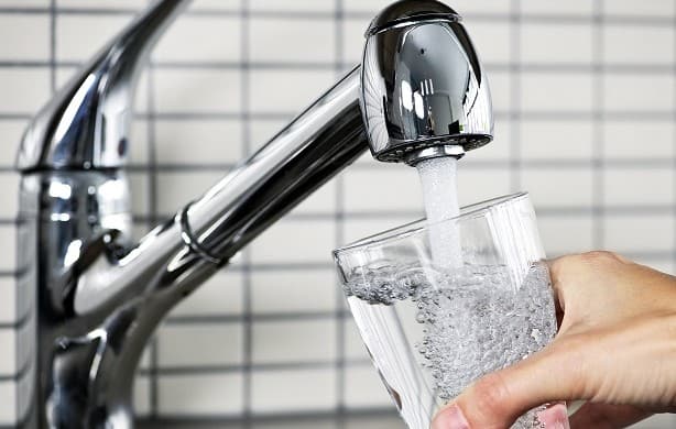 Close-up of a hand holding a glass being filled with water from a modern silver faucet against a tiled background.