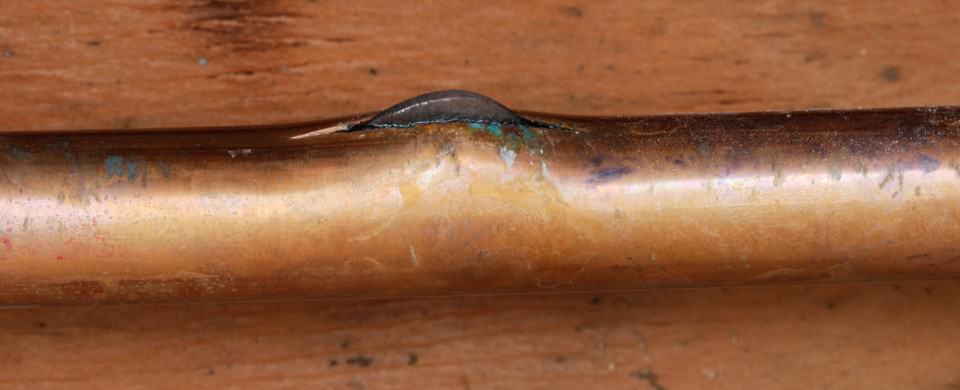 A copper pipe with a visible bulge and discolored edges, resting on a wooden surface.