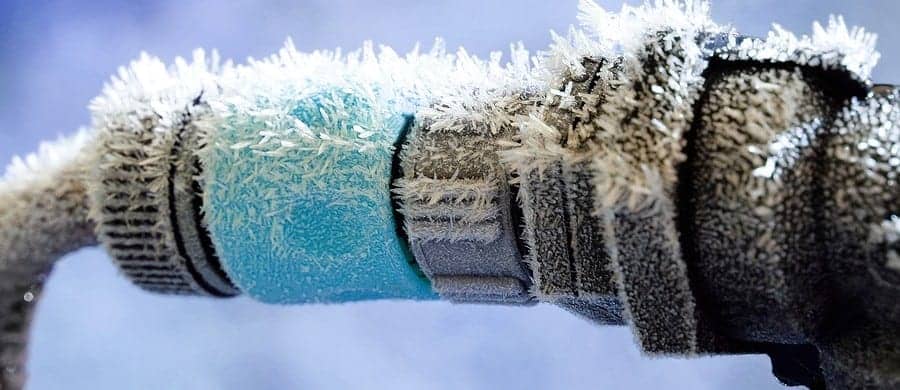 A close-up of a frozen pipe covered in ice crystals, showing a blue insulated section. The background is blurred, highlighting the icy texture.