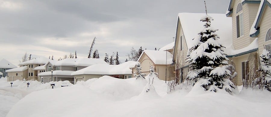 Snow-covered suburban neighborhood with houses partially obscured by snowbanks and snow-laden trees under a cloudy sky.